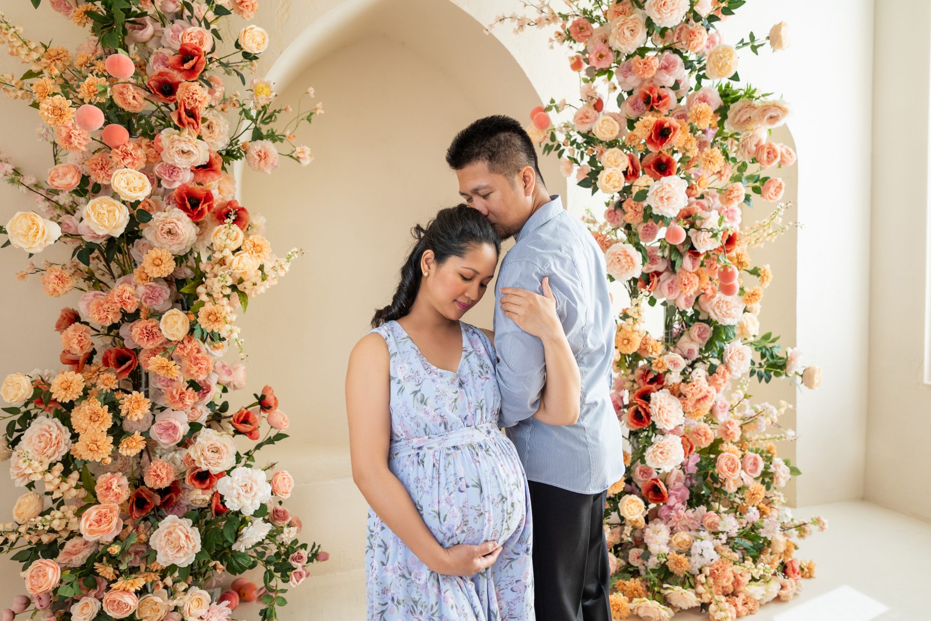 An elegant indoor pregnancy photoshoot in Milton featuring a couple in front of a beautiful floral archway in a light-filled studio.