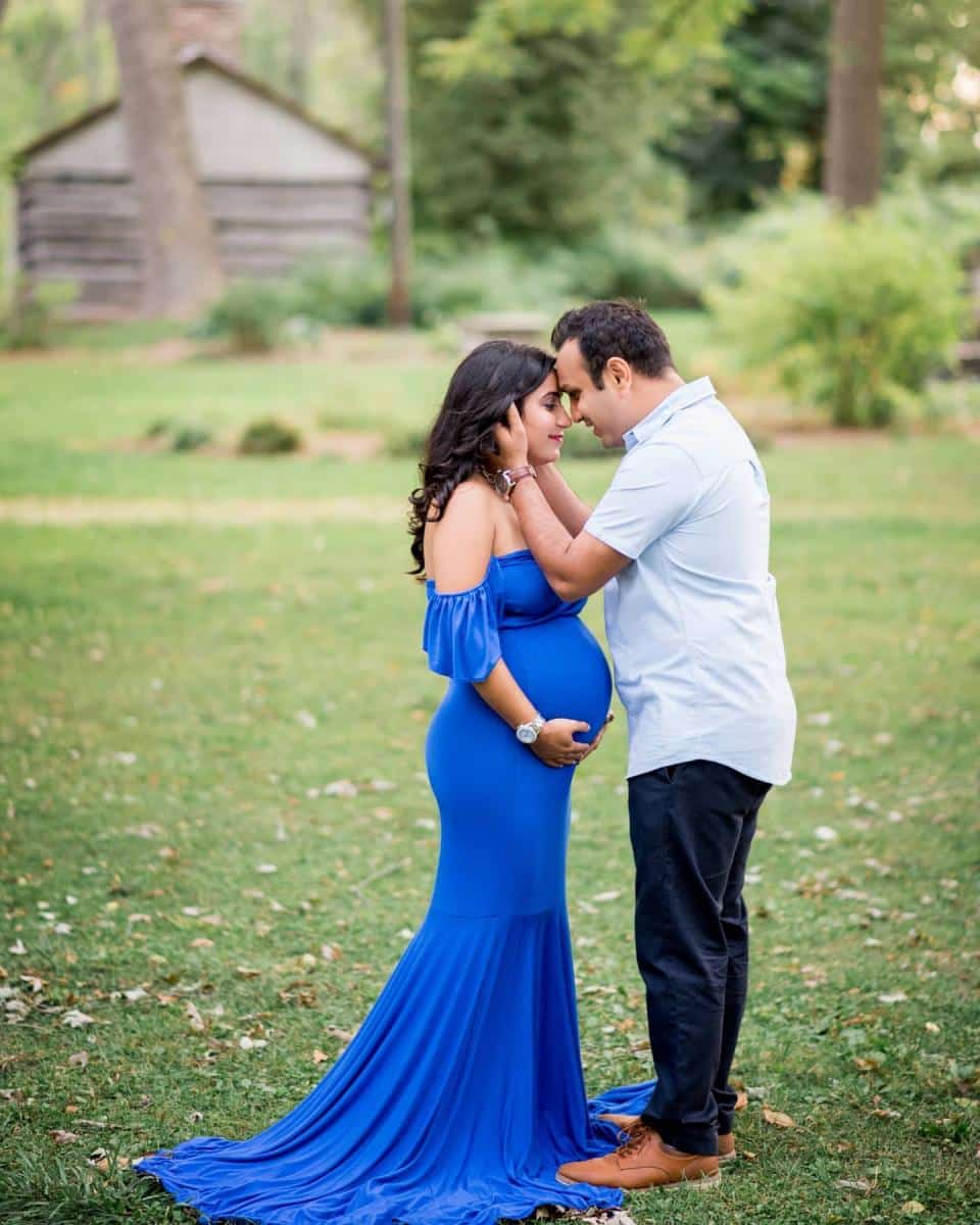 A tender moment between an expectant couple in a wooded area, with the woman wearing a flowing blue dress for her pregnancy photoshoot Brampton.