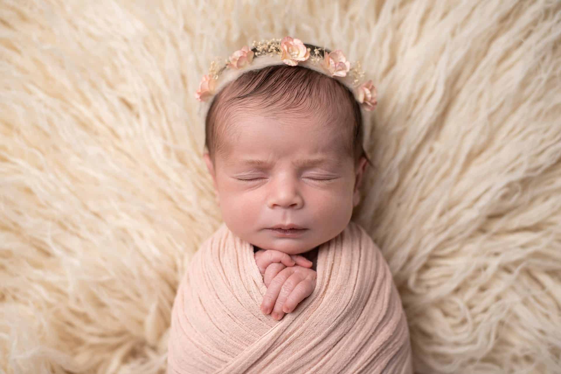Serene newborn baby girl sleeping on a fluffy white blanket, wrapped in pink with a floral headband, by a top newborn photographer Mississauga