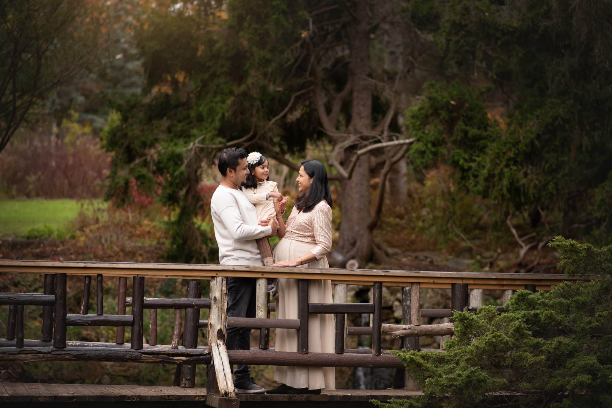 A scenic pregnancy photoshoot in Vaughan of a couple standing on a wooden bridge in a wooded area, sharing a quiet moment.
