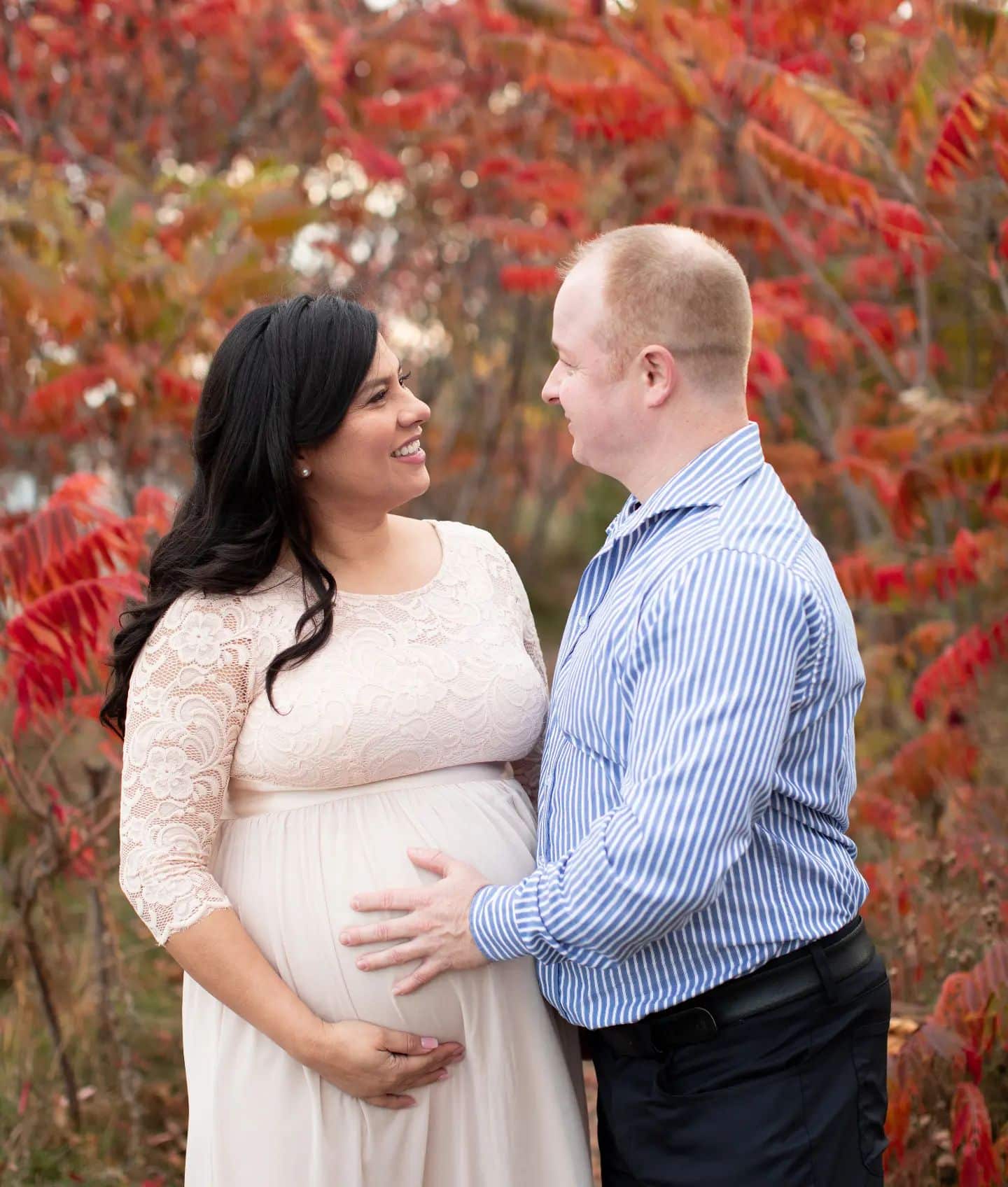 An outdoor maternity photography session in Oakville featuring a couple posing against vibrant red autumn leaves.