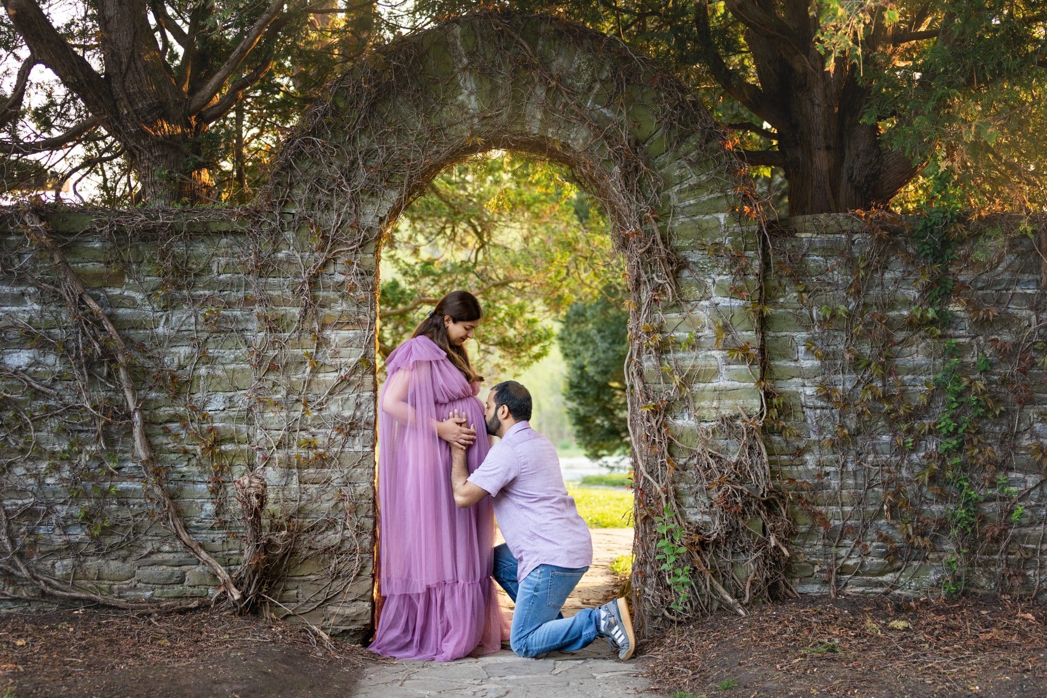 A creative maternity photography session in Oakville featuring a man kneeling to kiss the pregnant woman's belly under a stone archway.
