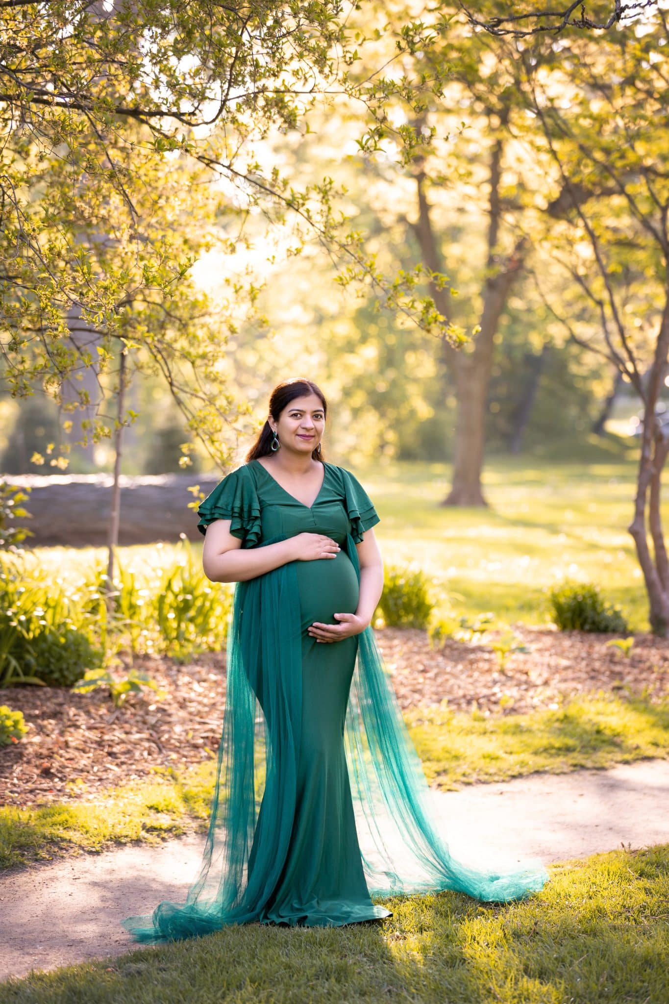 An ethereal pregnancy photoshoot in Mississauga showing a woman in a flowing emerald green gown standing in a sun-drenched park.