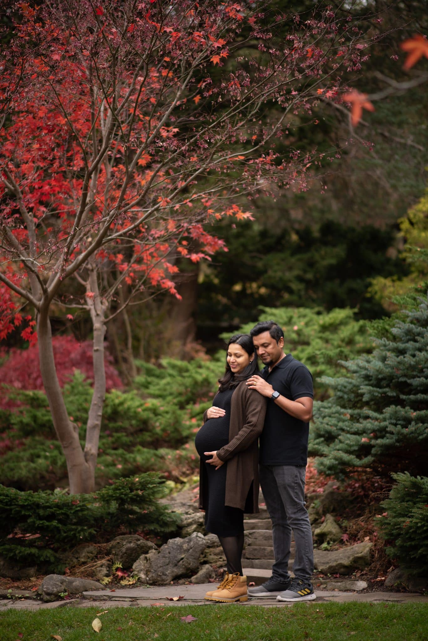 An autumn-themed maternity photo session in Richmond Hill featuring a couple standing under a tree with vibrant red fall leaves.