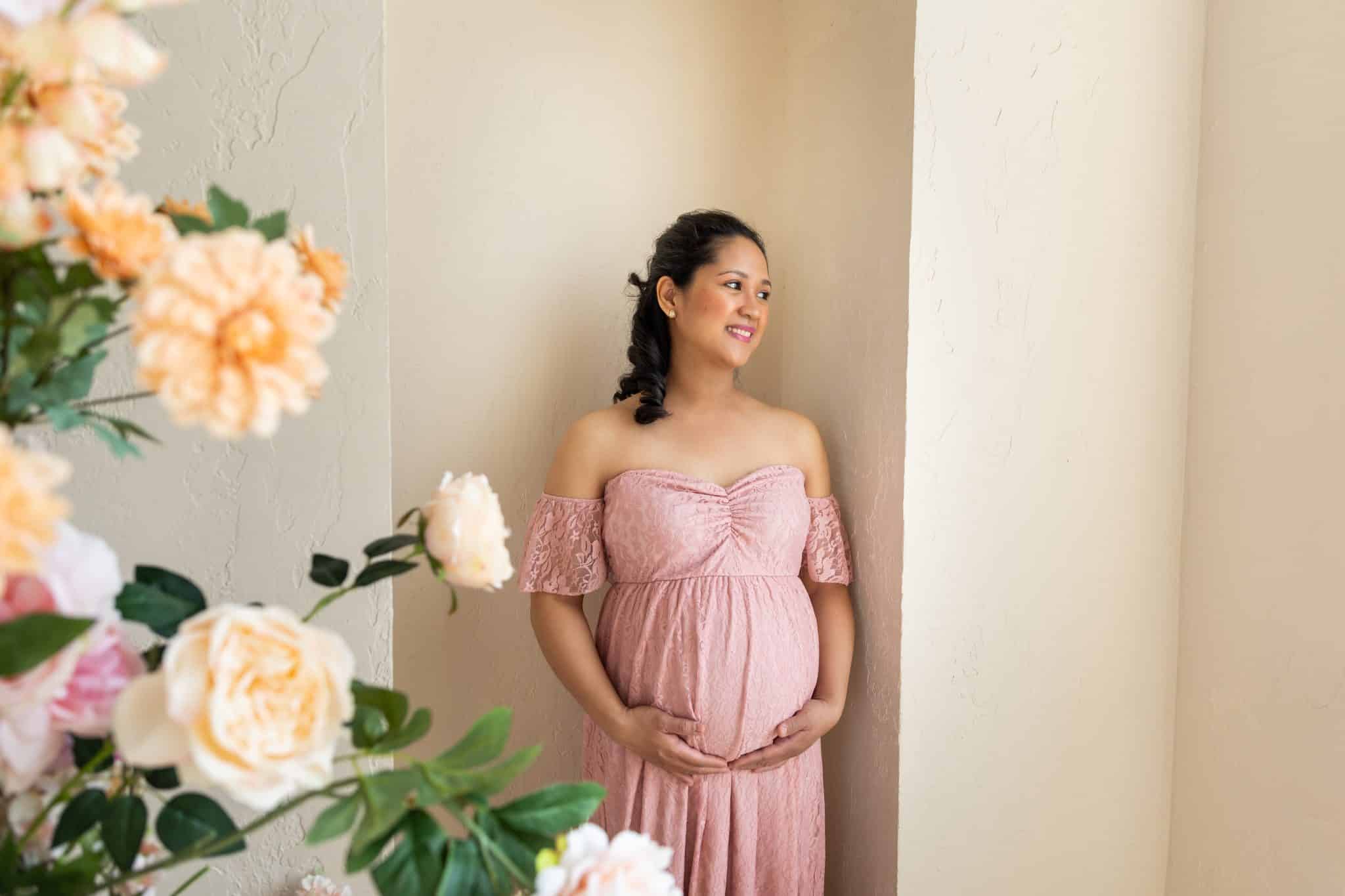A soft and elegant maternity photography session in Toronto featuring a woman in a pink lace dress posing next to large, peach-colored flowers in a studio.