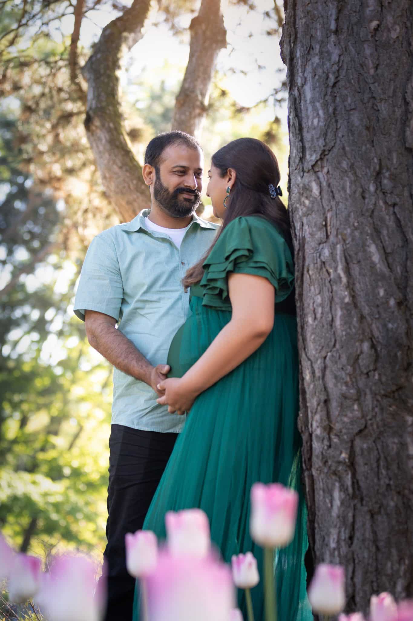 A lush green maternity photography session in Toronto featuring a pregnant woman in a green dress leaning against a tree with pink flowers in the foreground.