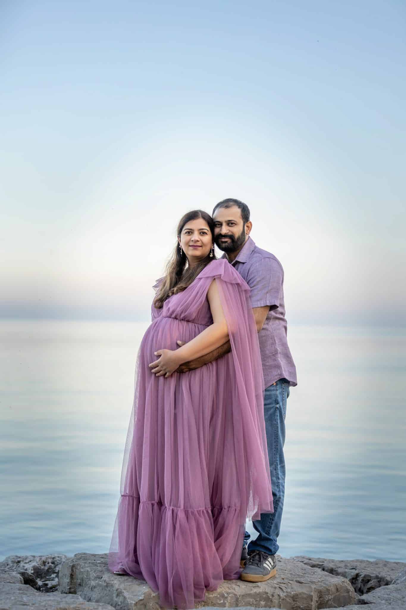 A stunning sunset maternity photography session in Burlington on a rocky beach with a woman in a flowing purple dress.