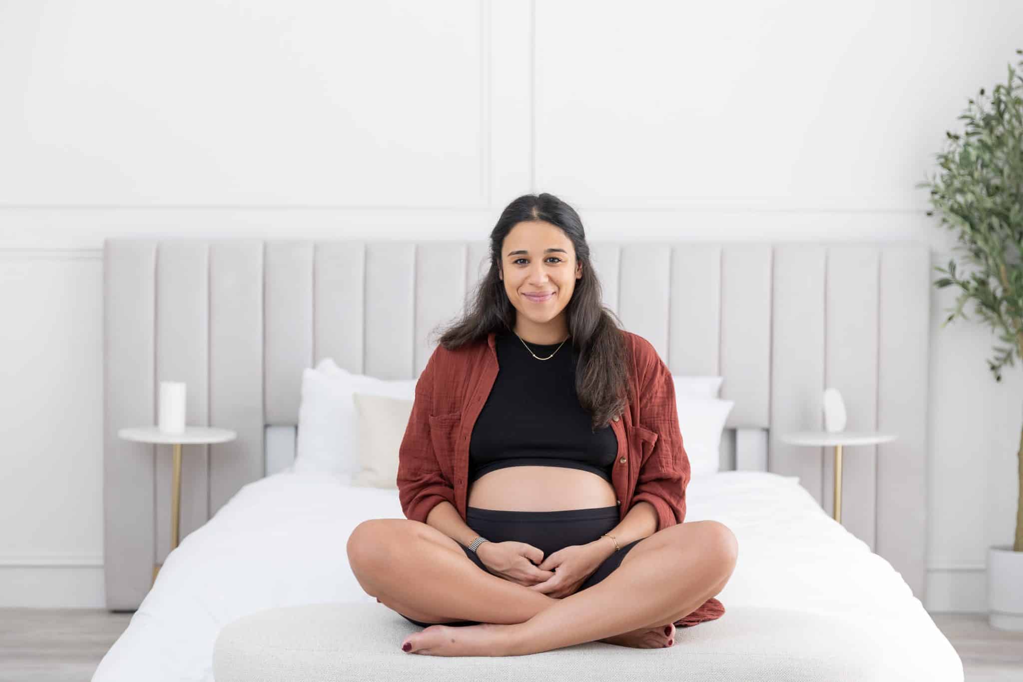 A lifestyle maternity photography session in Toronto featuring a pregnant woman sitting on a white bed, gently cradling her baby bump.