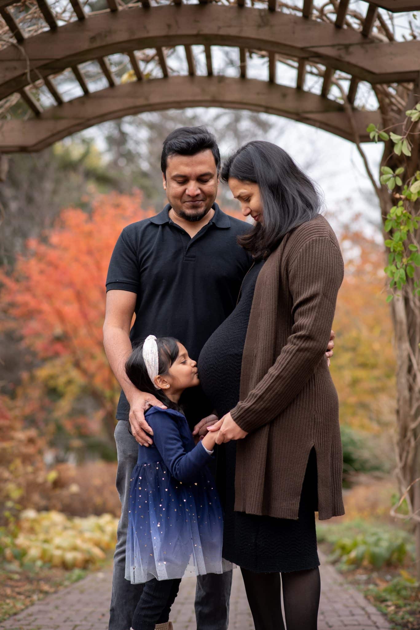 A family-focused maternity photography session in Burlington showing a couple with their young daughter, all posing together outdoors in late autumn.