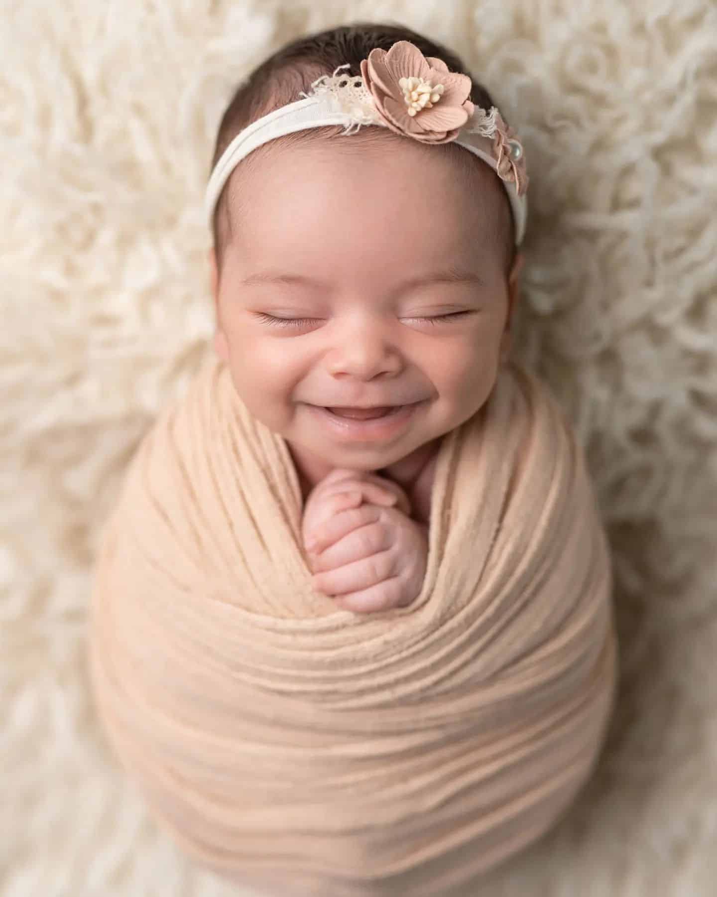 Smiling newborn baby girl wrapped in peach with a delicate headband, photographed by the best newborn photographer Mississauga offering affordable newborn photography Mississauga.