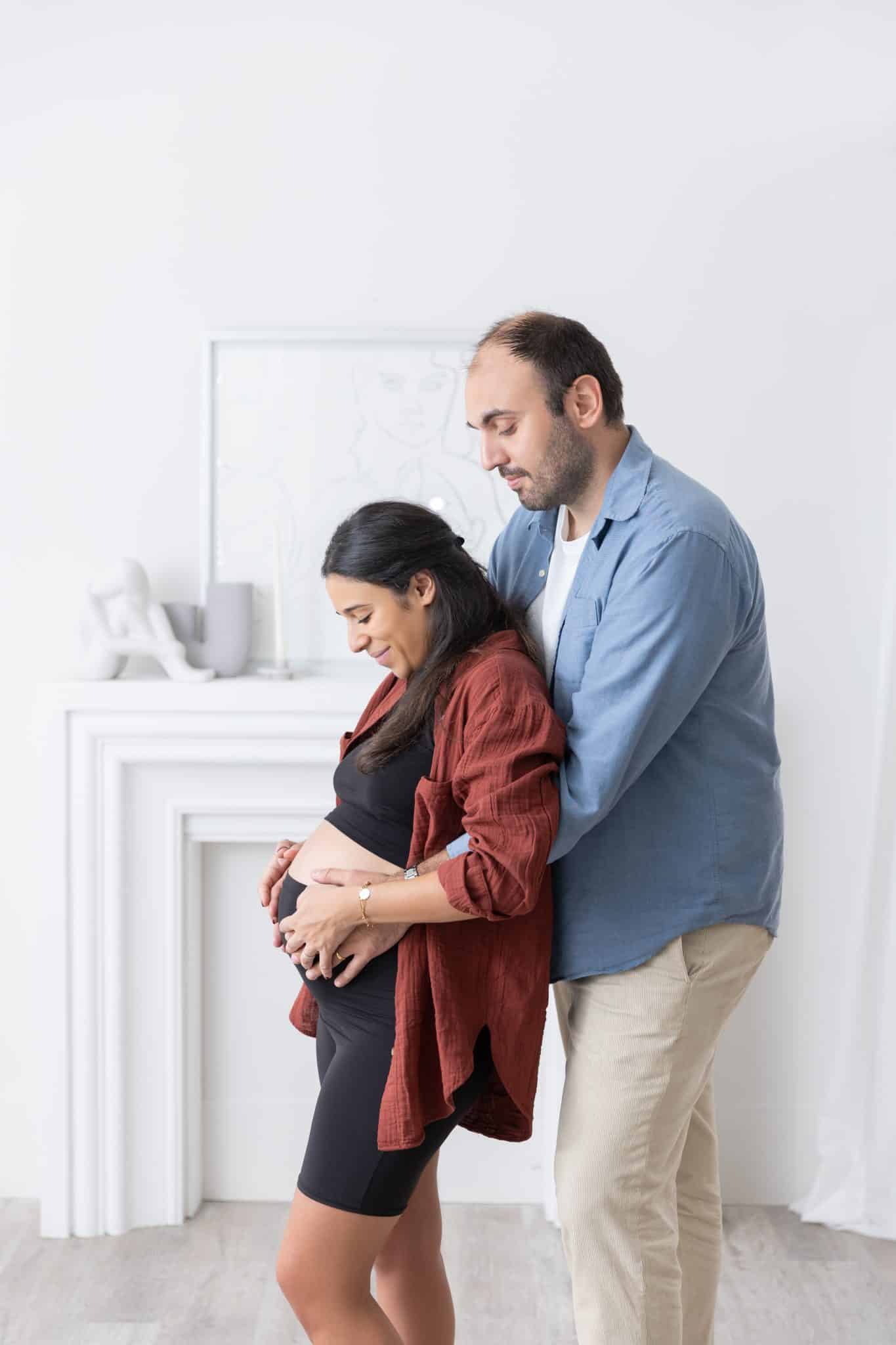 A tender pregnancy photoshoot in Mississauga showing a couple in a bright studio, looking down lovingly at the woman's belly.