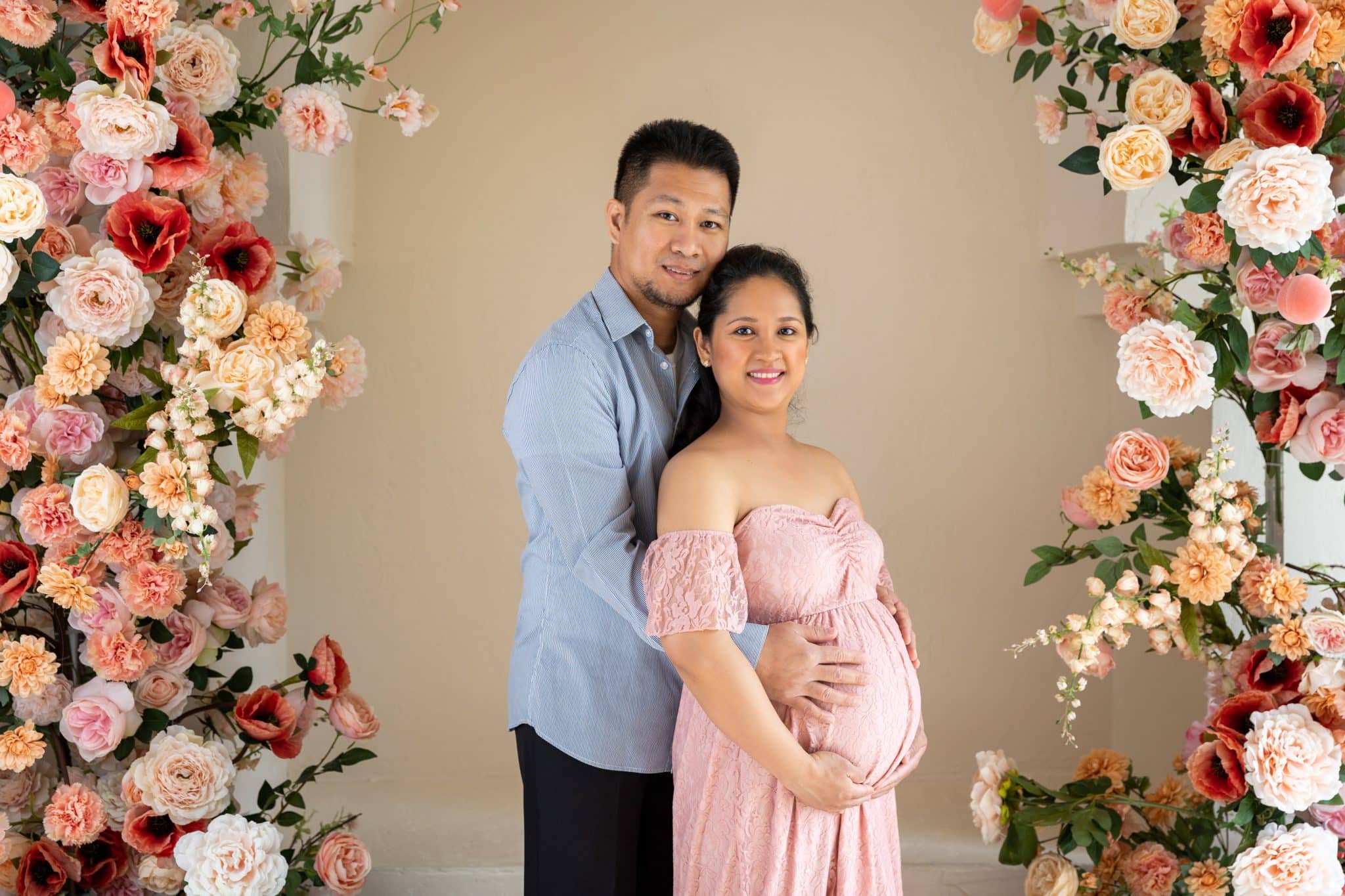 A beautiful studio maternity photo session in Brampton featuring a couple posing together within a vibrant floral archway, celebrating their pregnancy.