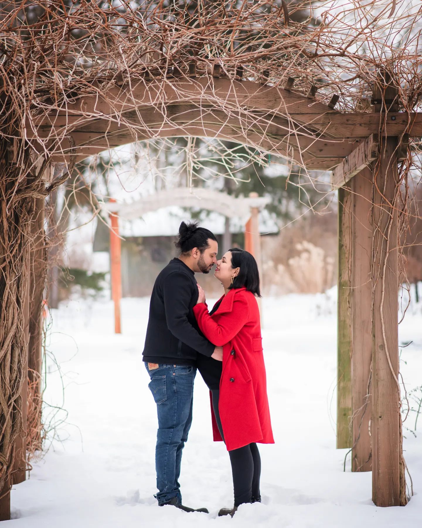 A romantic winter maternity photo session in Brampton with a couple kissing under a wooden pergola in a snowy outdoor setting.